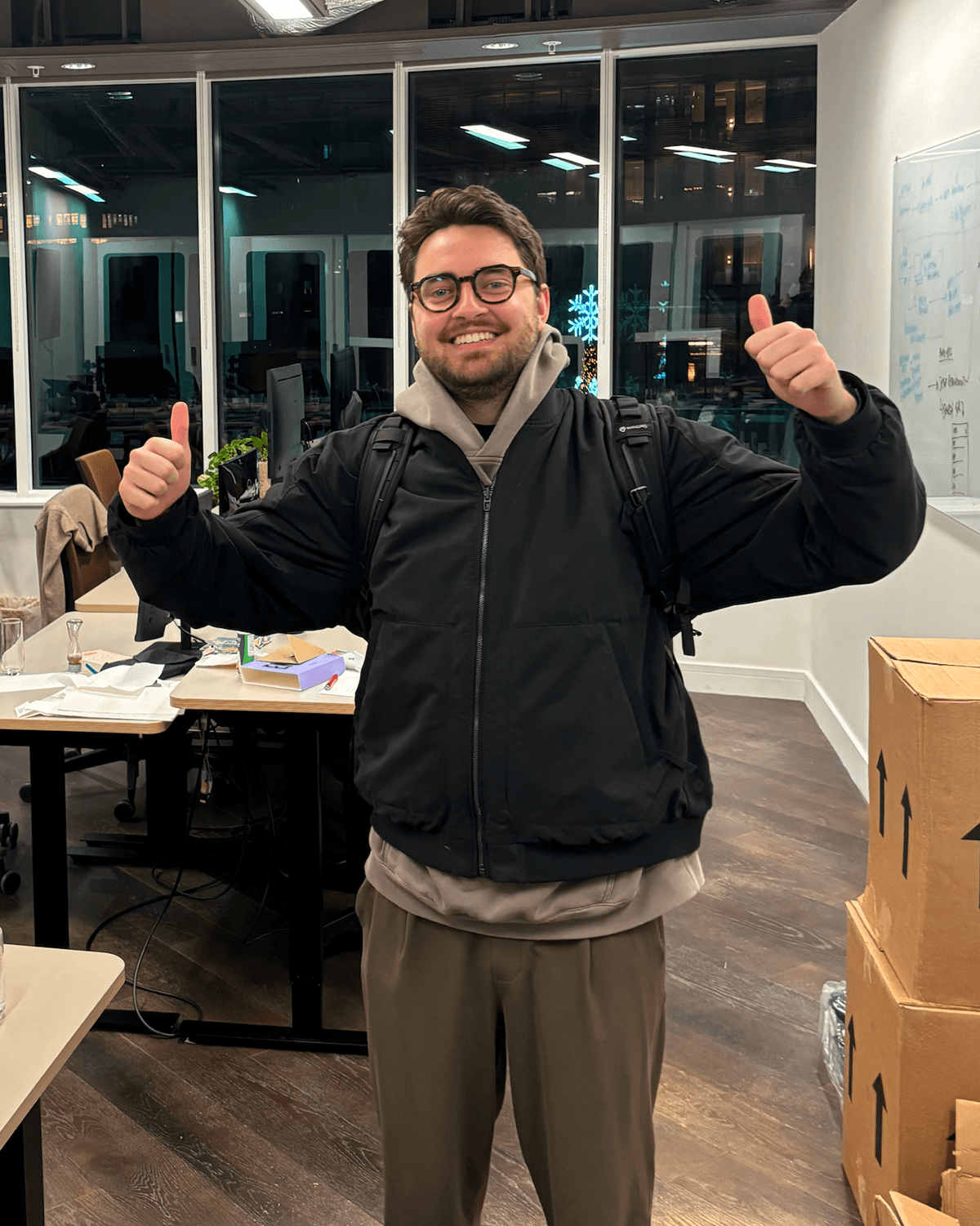 Alex Dunlop smiling and giving two thumbs up in an office workspace while wearing a black jacket, hoodie, and backpack.